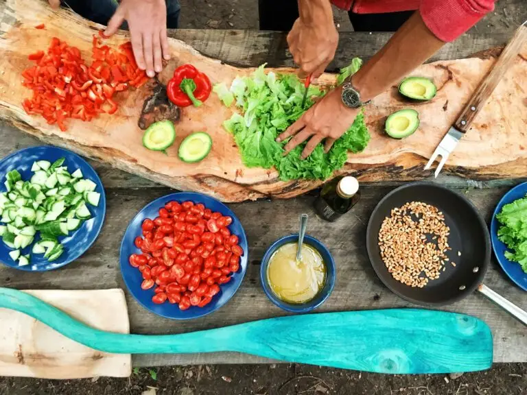 top view of 2 people preparing a salad, with lettuce, bell peppers and avocado on a beautiful wooden cutting board, and in separate blue bowls bits of cucumber, tomato, dressing and a pan with roasted pine nuts.