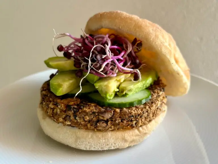 A plant-based burger featuring a hearty veggie patty, sliced cucumber, avocado, and vibrant purple sprouts, served in a soft bun on a white plate.