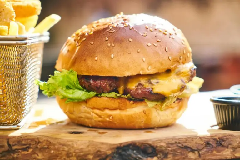 A close-up of a juicy high cholesterol cheeseburger with lettuce, cheese, and a sesame seed bun, accompanied by a basket of crispy French fries in the background.