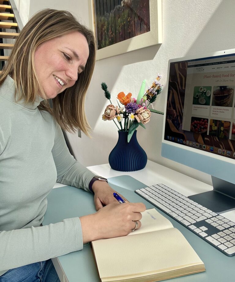 Dr. Sharon Facey, a family doctor specializing in menopause and heart health, writing in a notebook while sitting at her desk. A computer screen displays her blog, ‘(Plant-Based) Food for Thought,’ featuring heart-healthy recipes and science-backed wellness tips.
