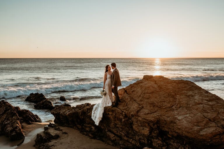 Bride and groom standing out on a large rock with the ocean sunset behind them during their Crystal Cove wedding