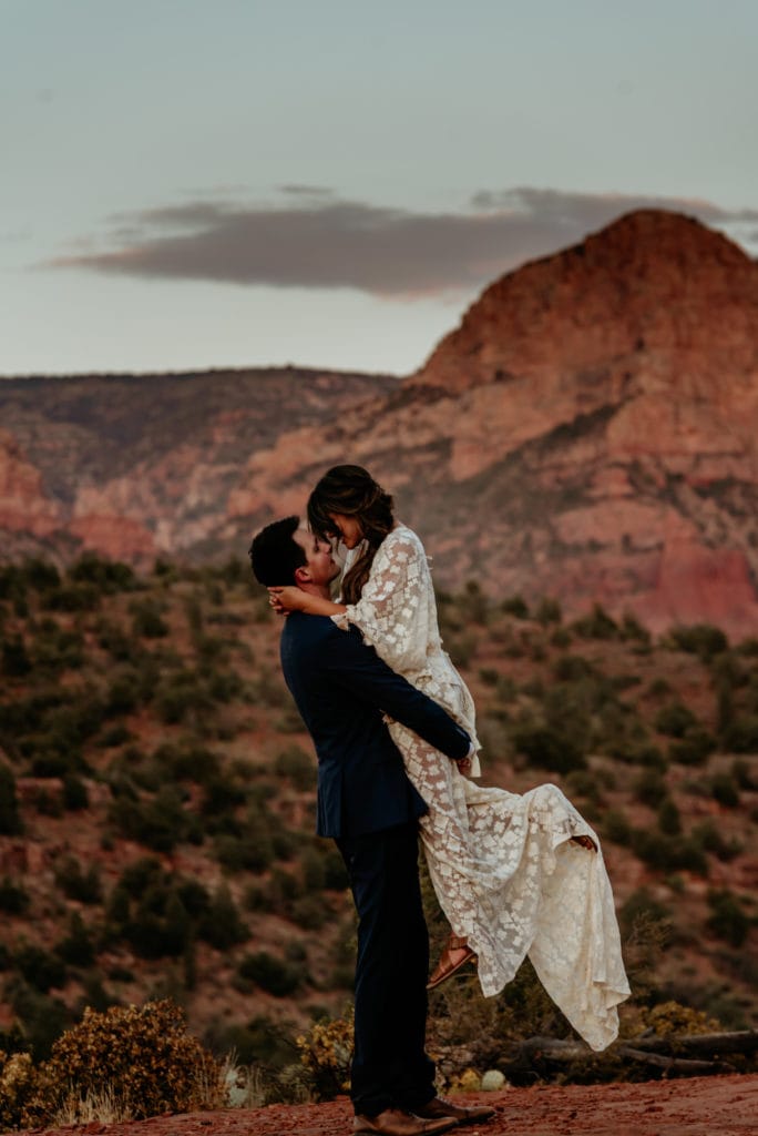 Husband lifts his wife in her long lace dress surrounded by Sedona red rocks at sunset
