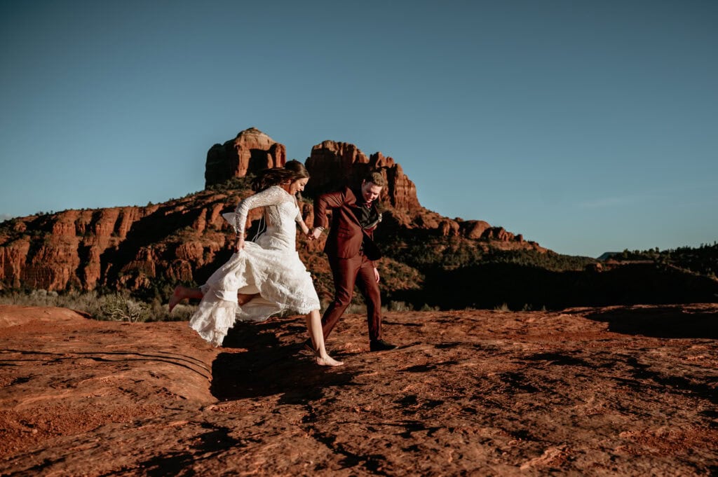 Barefoot bride leaps over a crack in the rocks with the help of her groom during  their Sedona elopement