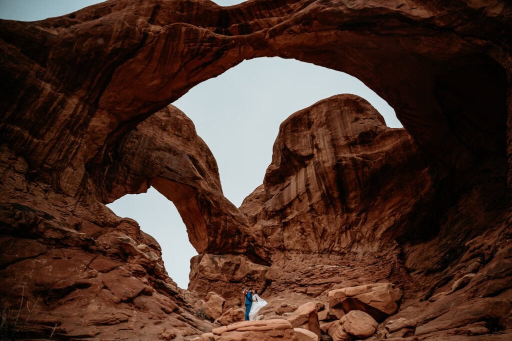 Couple standing on a boulder with double arch above them during their Moab elopement photos
