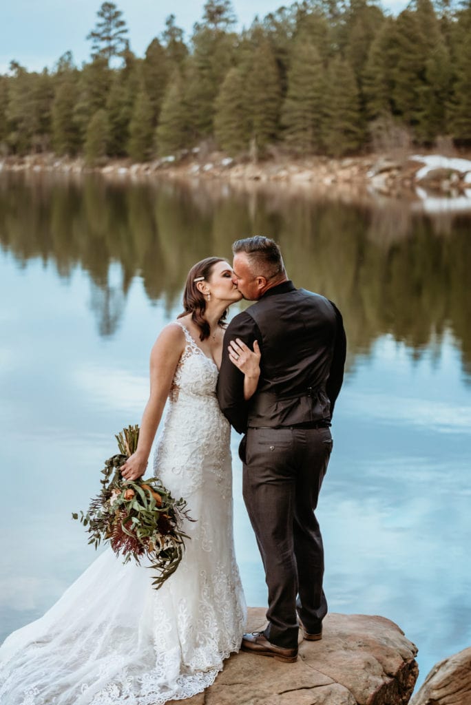 Elopement couple embrace for a kiss framed by the reflection of the tree lined winter lake