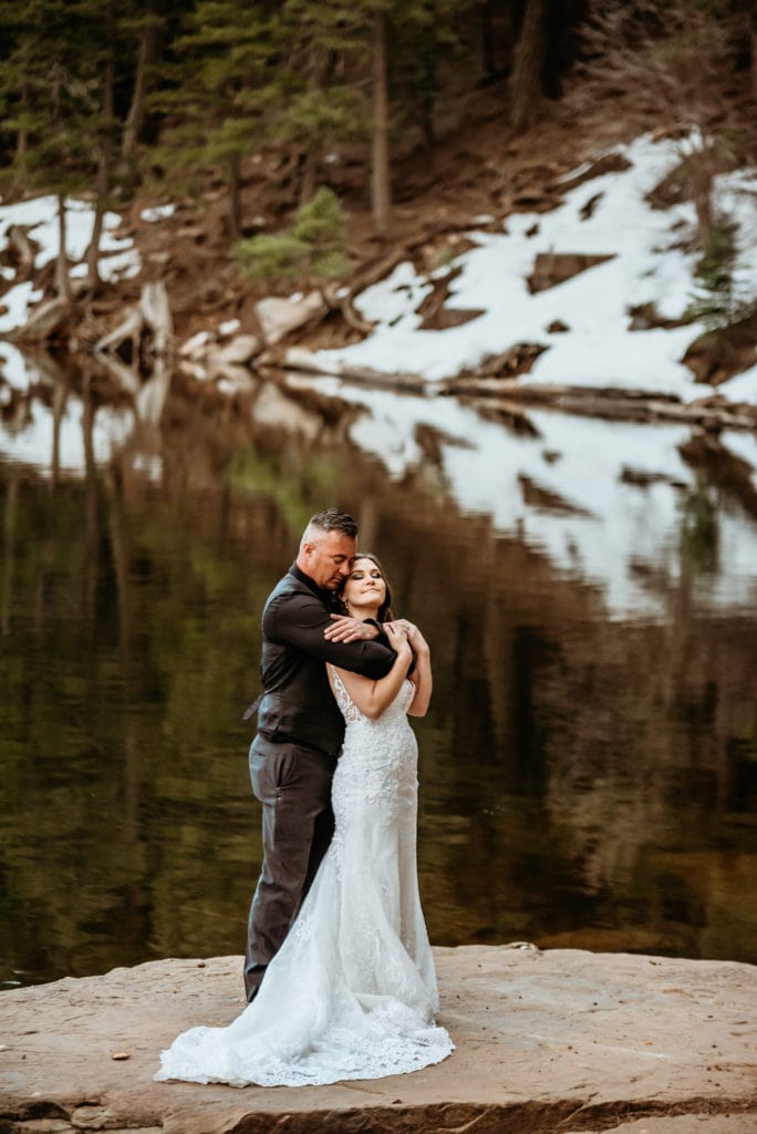 Groom holds his bride tight on the edge of a snow lined lake