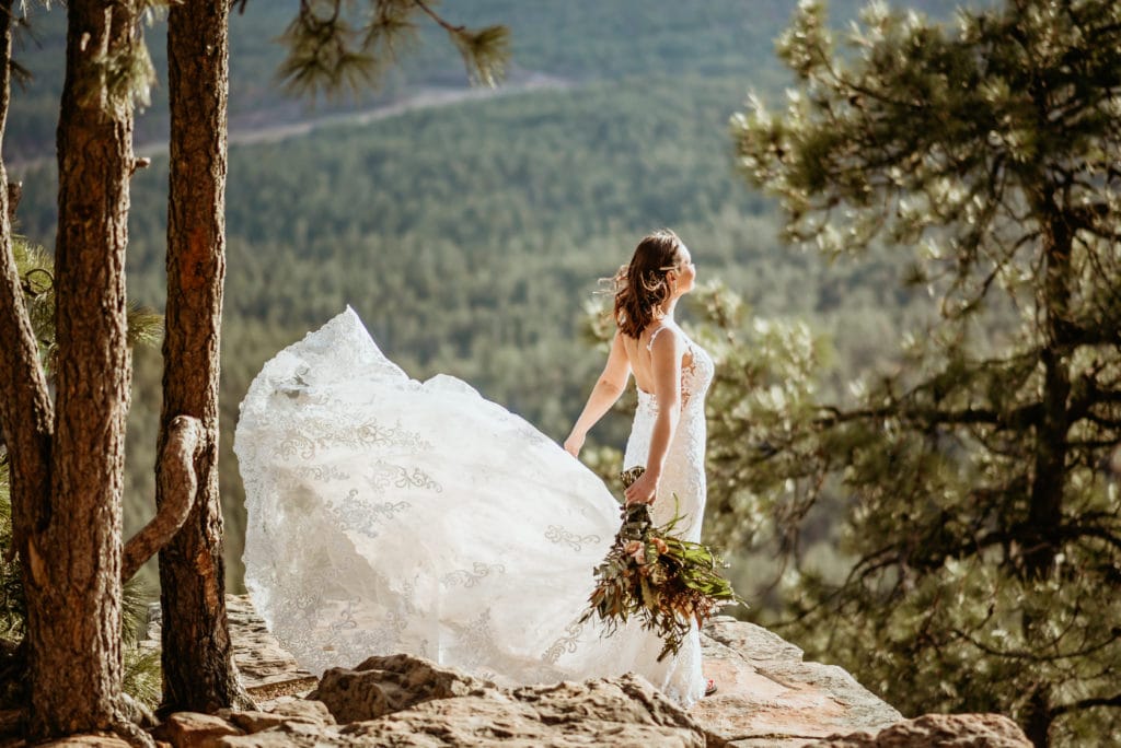 Bride's long wedding dress train blowing in the wind as she stands at the edge of the world on a cliff overlooking the Mogollon Rim