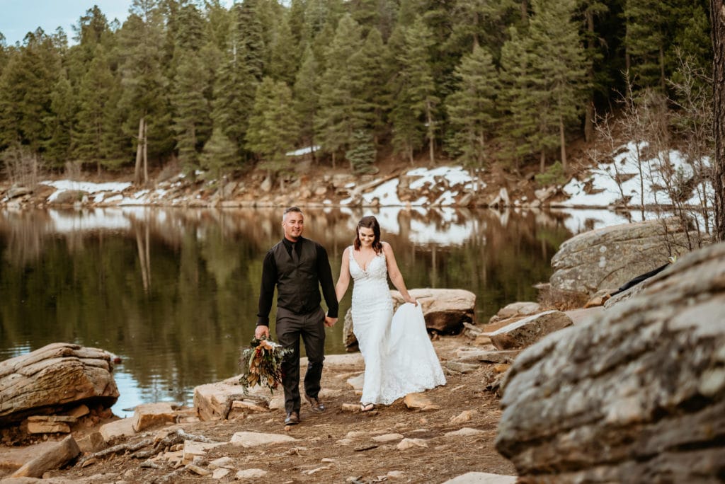 Patches of snow and evergreens line the bank of a wintery lake as bride and groom walk hand in hand