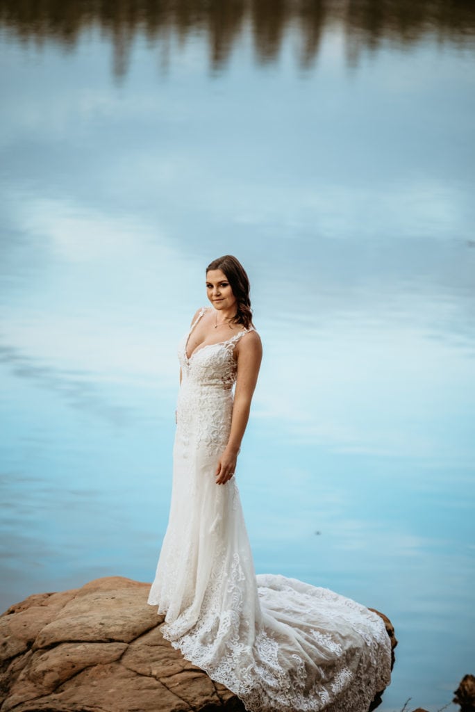 Stunning bride on a stone with light blue sky reflecting in the lake behind her