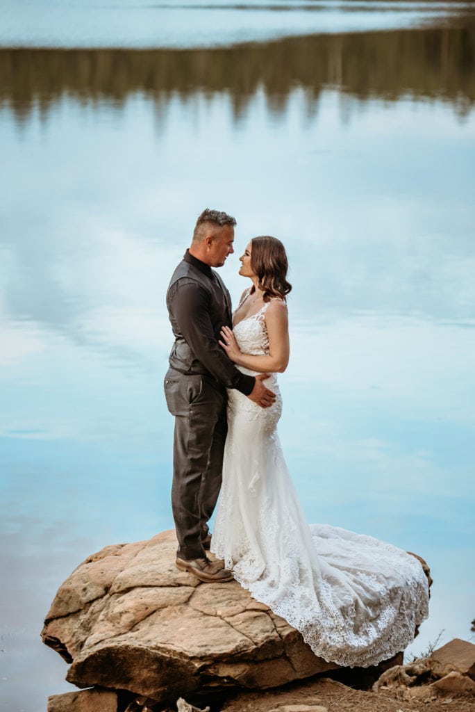 Bride and groom embrace, framed in winter lake reflecting the sky and pine trees