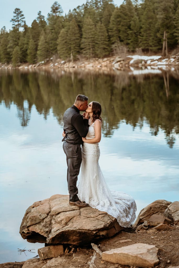 Groom lifts bride's chin as he draws her in for a kiss next to a beautiful winter lake during their Arizona elopement