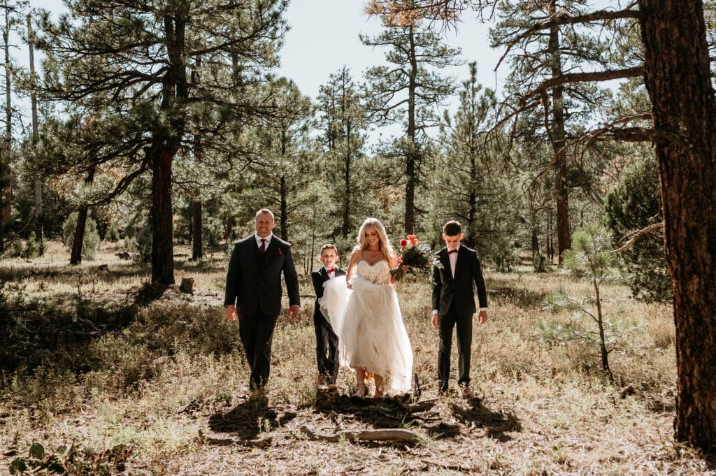 bride and groom walk through pine trees as sons help carry her dress during their elopement with family