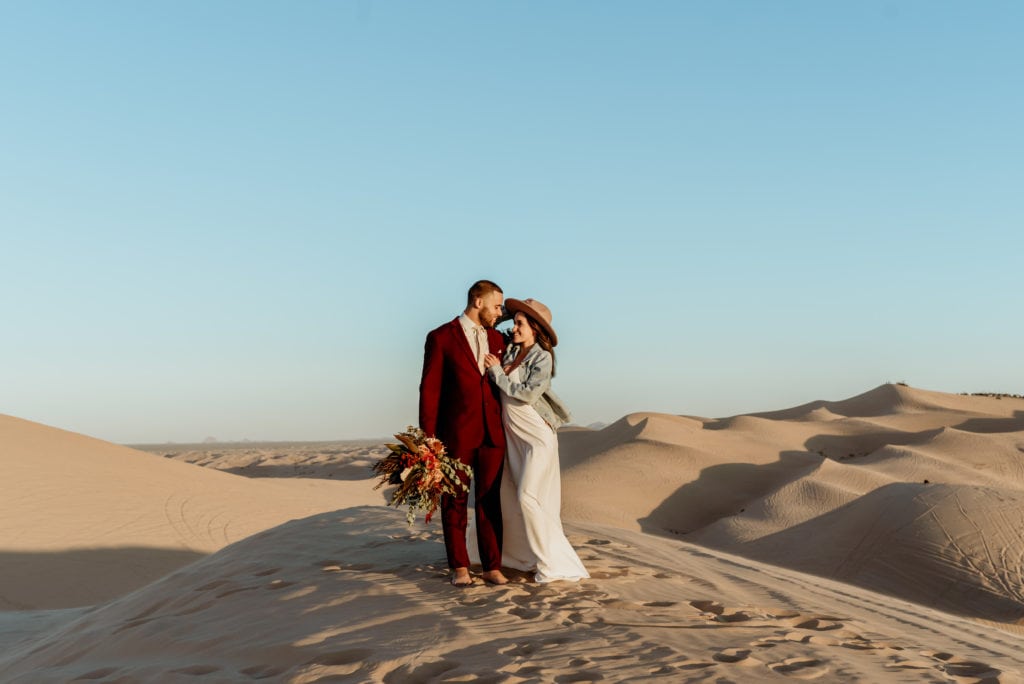 Bride holds her hat to keep it from blowing away while embracing her husband on top of waves of golden sand