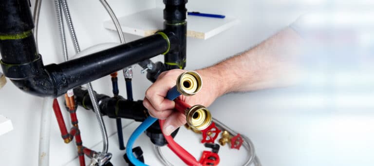 A person holds red and blue flexible pipes beside plumbing fixtures under a sink.