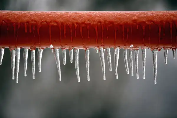 A red pipe with icicles hanging from it, set against a blurred gray background.