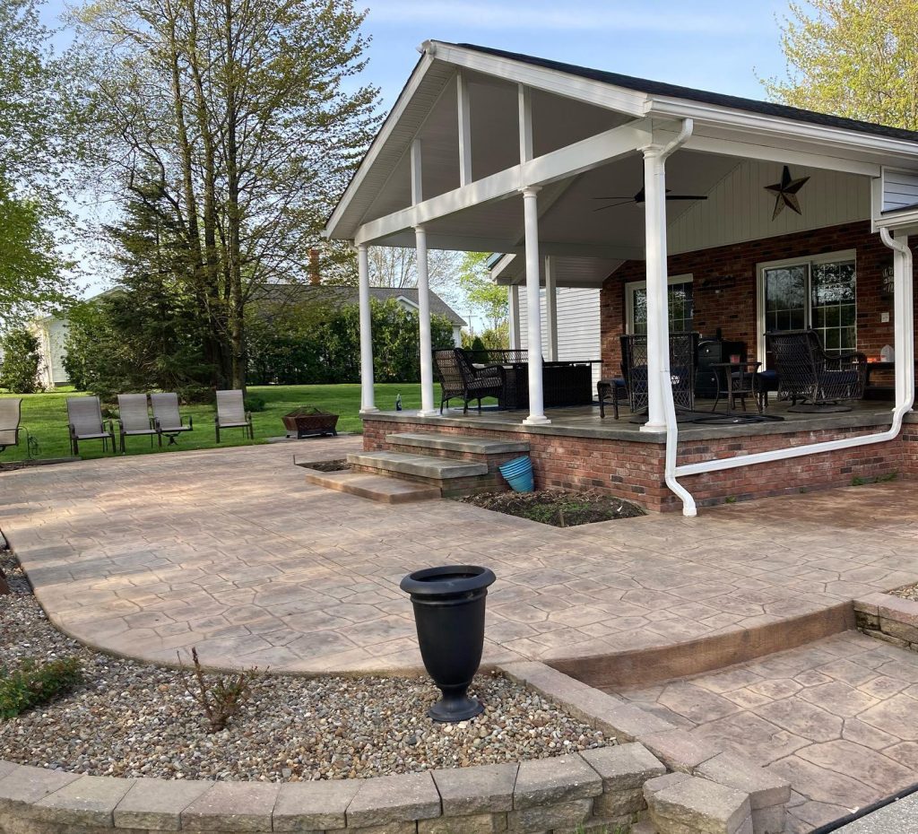 A backyard patio with quality concrete construction features stamped paving, a raised covered porch, outdoor chairs, and a large yard with trees and grass. A black planter sits on the patio, and a fire pit with chairs is in the background.