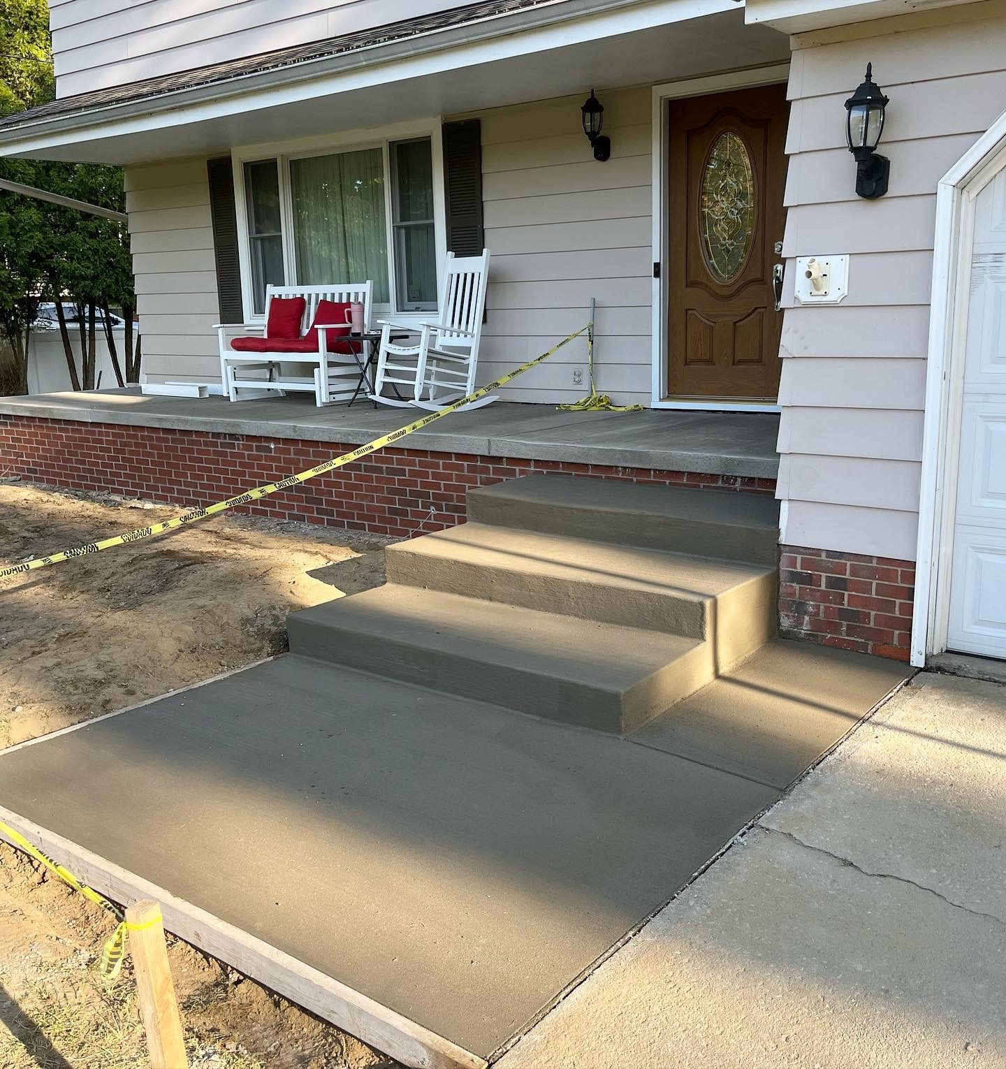 Fresh concrete construction steps and walkway lead to a front porch with beige siding and a wooden door. Yellow caution tape marks the area, while a white rocking chair and red bench sit invitingly on the porch.