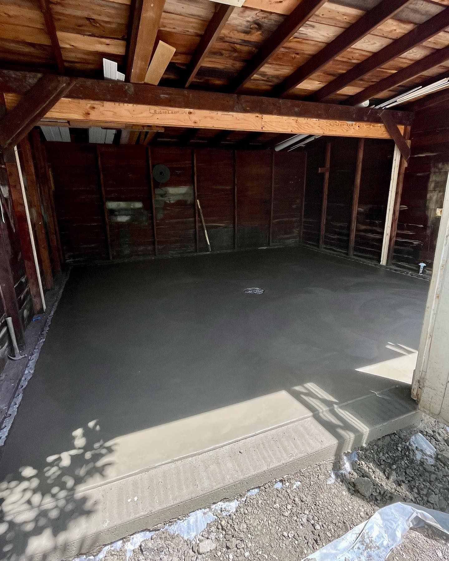 A garage with exposed wooden beams and walls, showcasing a freshly poured and smoothed concrete construction floor. Sunlight streams in from the entrance, casting shadows on the new surface.