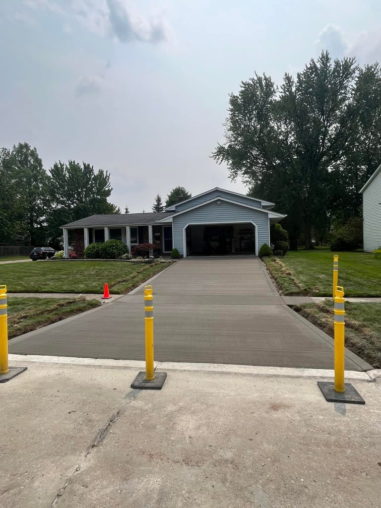 A freshly poured concrete driveway, the result of expert concrete construction, leads up to a white house with an open garage. Yellow barriers and a single orange cone block access, and the surrounding lawn is neatly trimmed.