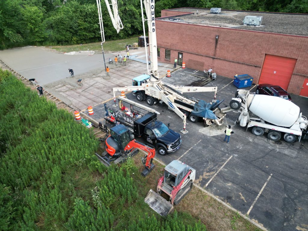 Aerial view of a construction site where a construction company is pouring concrete. Multiple trucks, paving equipment, and a red-brick building with a red door and dumpster are visible, surrounded by trees and vegetation.