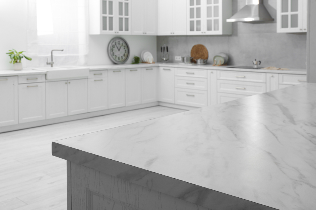 Modern kitchen with white cabinets, a marble countertop in the foreground, stainless steel appliances, and minimalist decor built by a leading construction company. Natural light enters through a window with sheer curtains.