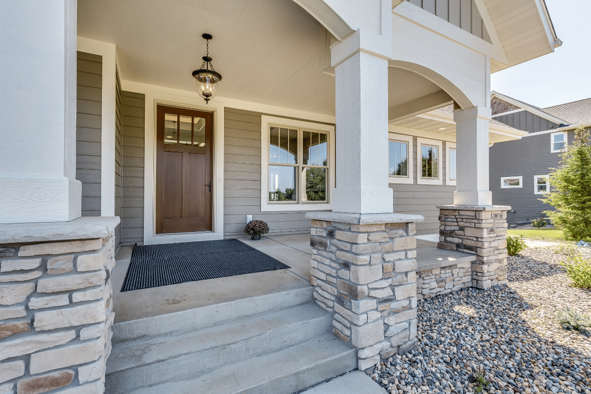 Front porch of a modern house with stone pillars, gray siding, a brown wooden door, large windows, a hanging lantern, concrete steps built with quality concrete construction, and a gravel landscaping area.