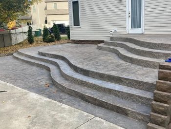 A modern backyard patio built by a trusted construction company features wide, curved concrete steps leading up to a house with light siding. The stamped concrete pattern and landscaped edges blend seamlessly with lush greenery in the background.