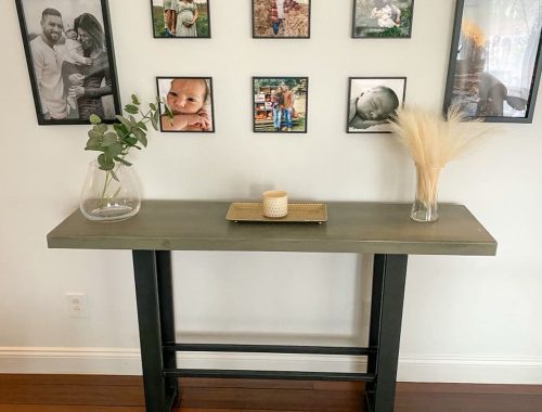 A modern console table with a glass vase of greenery, a tray with a candle, and dried wheat decor sits against a wall displaying family photos in black frames arranged in a grid, adding warmth to this concrete construction-inspired space.