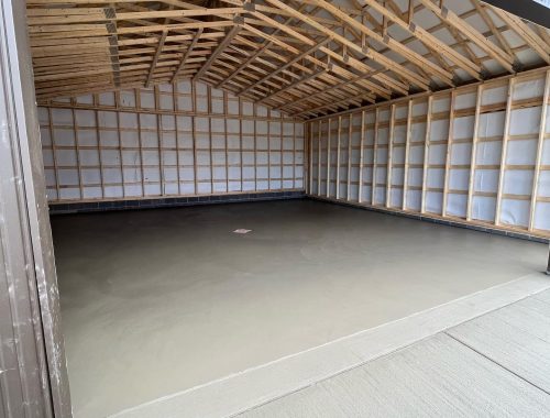 Interior view of a large, empty metal shed with unfinished wooden framing and a smooth, newly poured concrete floor—showcasing precise concrete construction. The structure features an open front and a high, peaked ceiling.