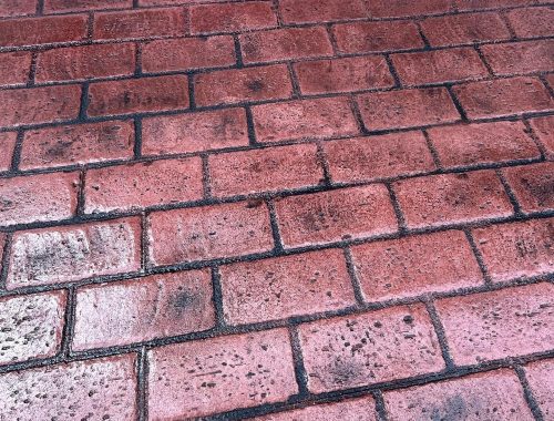 Red brick pavement with rectangular bricks arranged in a staggered paving pattern. The surface appears wet with scattered black specks and stains, likely remnants from an excavation or construction company project; a light blue wall tops the image.