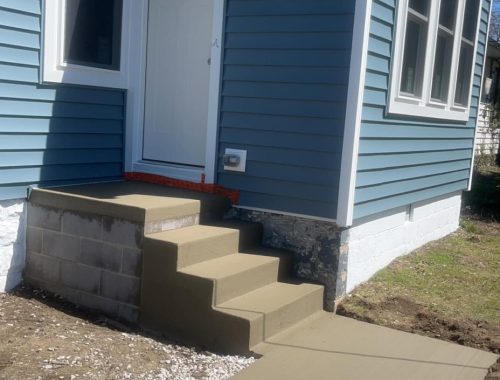 A blue house with white trim features a new concrete construction staircase and walkway leading to a white door. Gravel is spread at the base, soil surrounds the area, and the steps are unfinished—missing railing and paint.