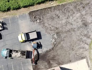 Aerial view of excavation and construction vehicles removing old asphalt from a parking lot, with exposed dirt covering part of the area and surrounding equipment visible as part of a concrete construction project.