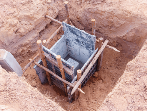 A concrete footing with wooden and metal formwork is set in a rectangular hole dug in the ground, prepared for a building’s foundation construction by an expert construction company.