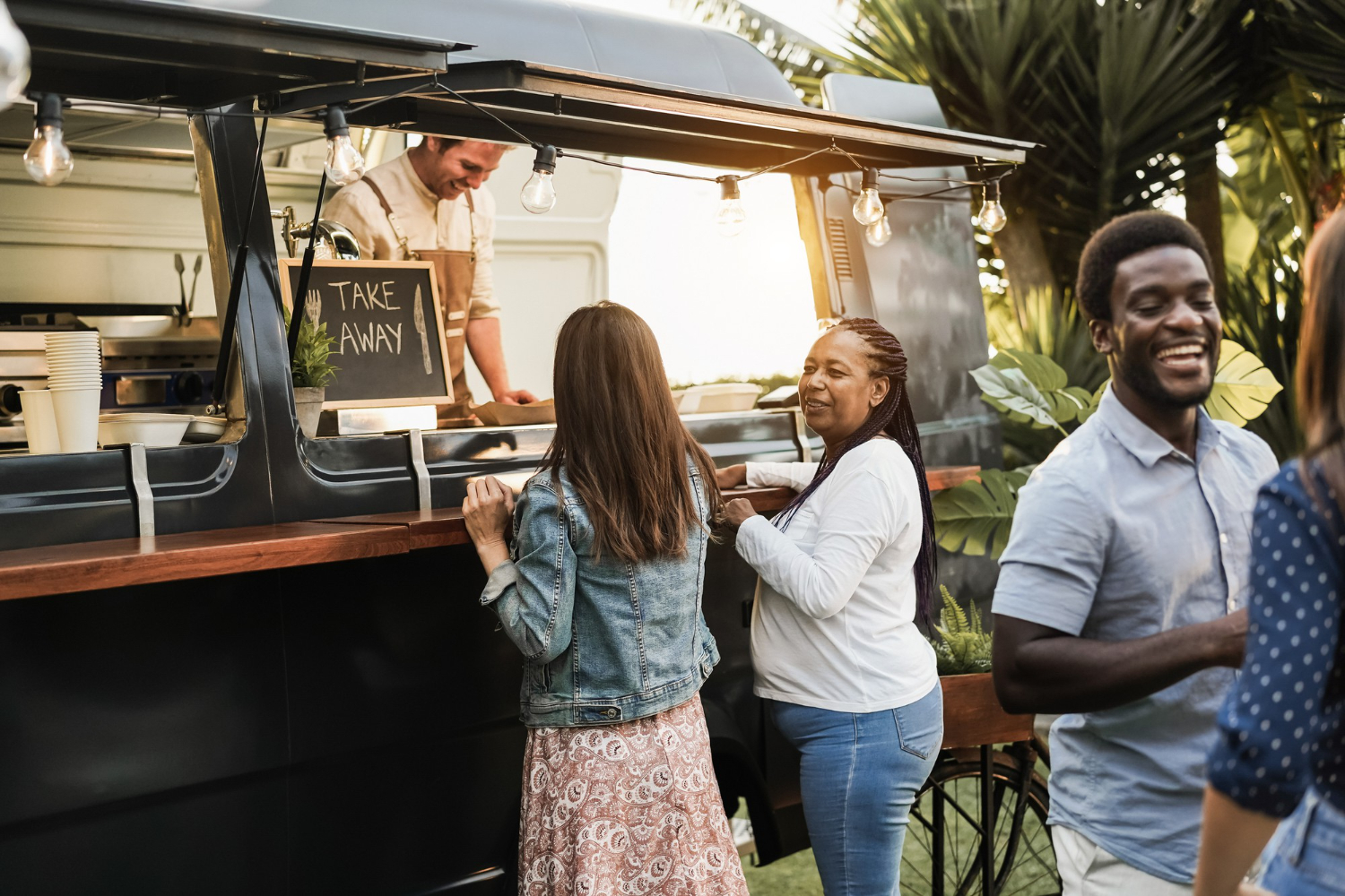 Two women stand at a food truck ordering window, ordering food and talking to each other.