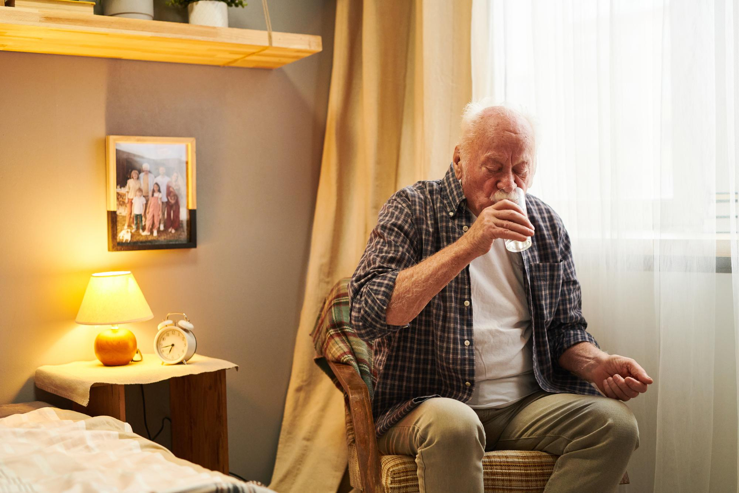 An older man sits in a chair in his bedroom and takes a sip of water from a glass as he takes his morning medication.