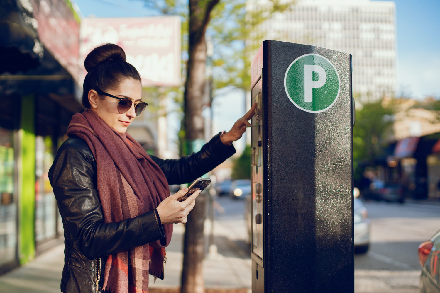 A young woman stands at a parking payment box, marked with a white letter P in a green circle. She's holding a smart phone and inserting her card into the payment box.
