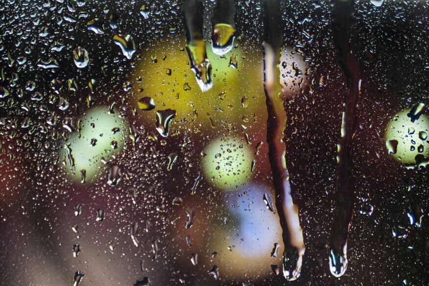 Water droplets from a rainstorm run down a clear window with blurry lights in the background.