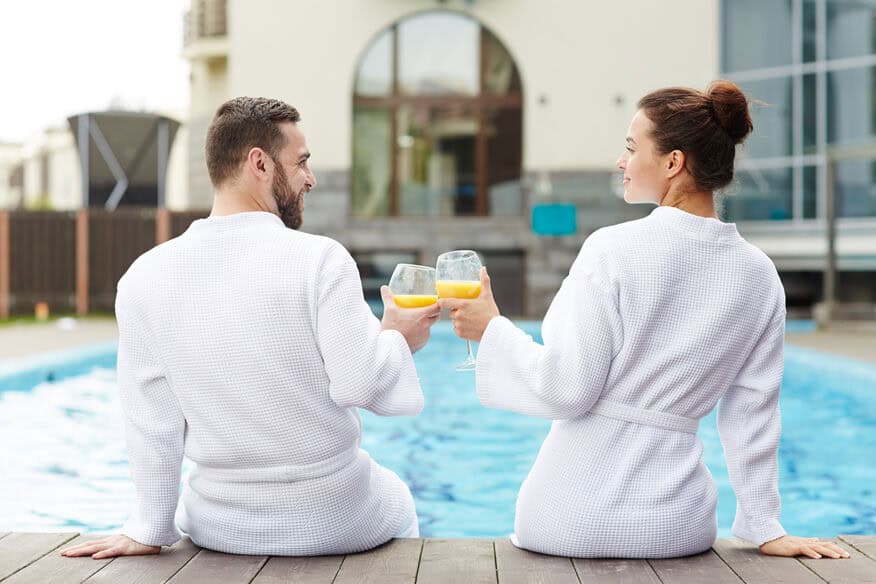 A couple sits at the edge of an outdoor hotel pool, wearing white hotel bathrobes and enjoying drinks together on a sunny day .