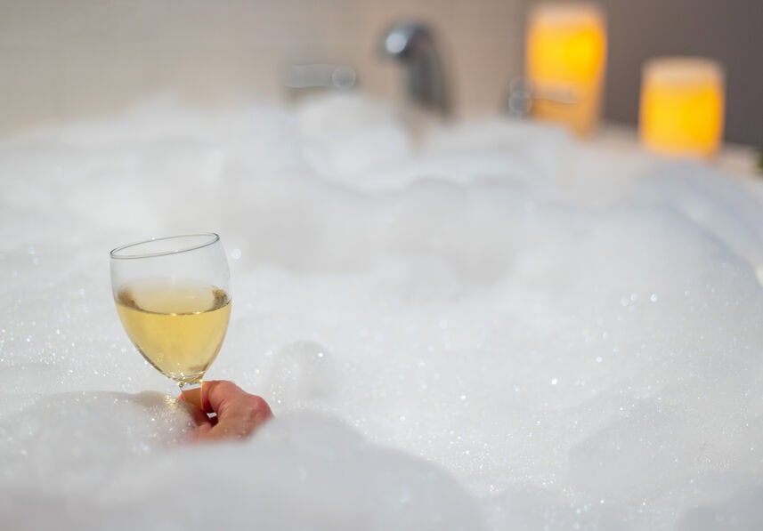 A woman’s hand holds a glass of white wine while relaxing in a bathtub full of soap bubbles.