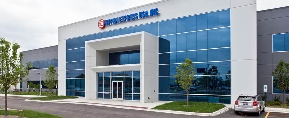 A white and grey office building behind a parking lot has blue mirror windows along the entrance wall and trees on each side of the front door. The sign above the entrance says “Nippon Express.”