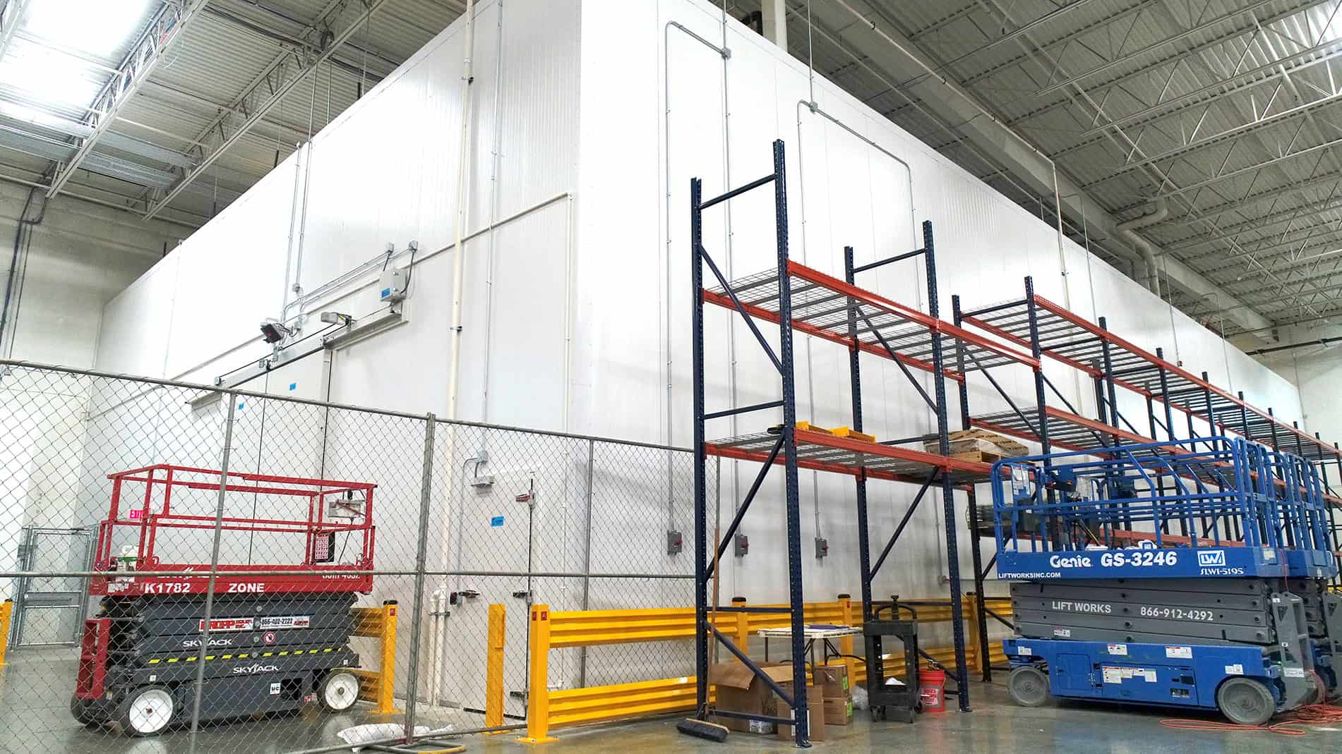 A Nippon Express warehouse has metal shelves and a forklift along the fenced outer walls of a GDP freezer and cooler.