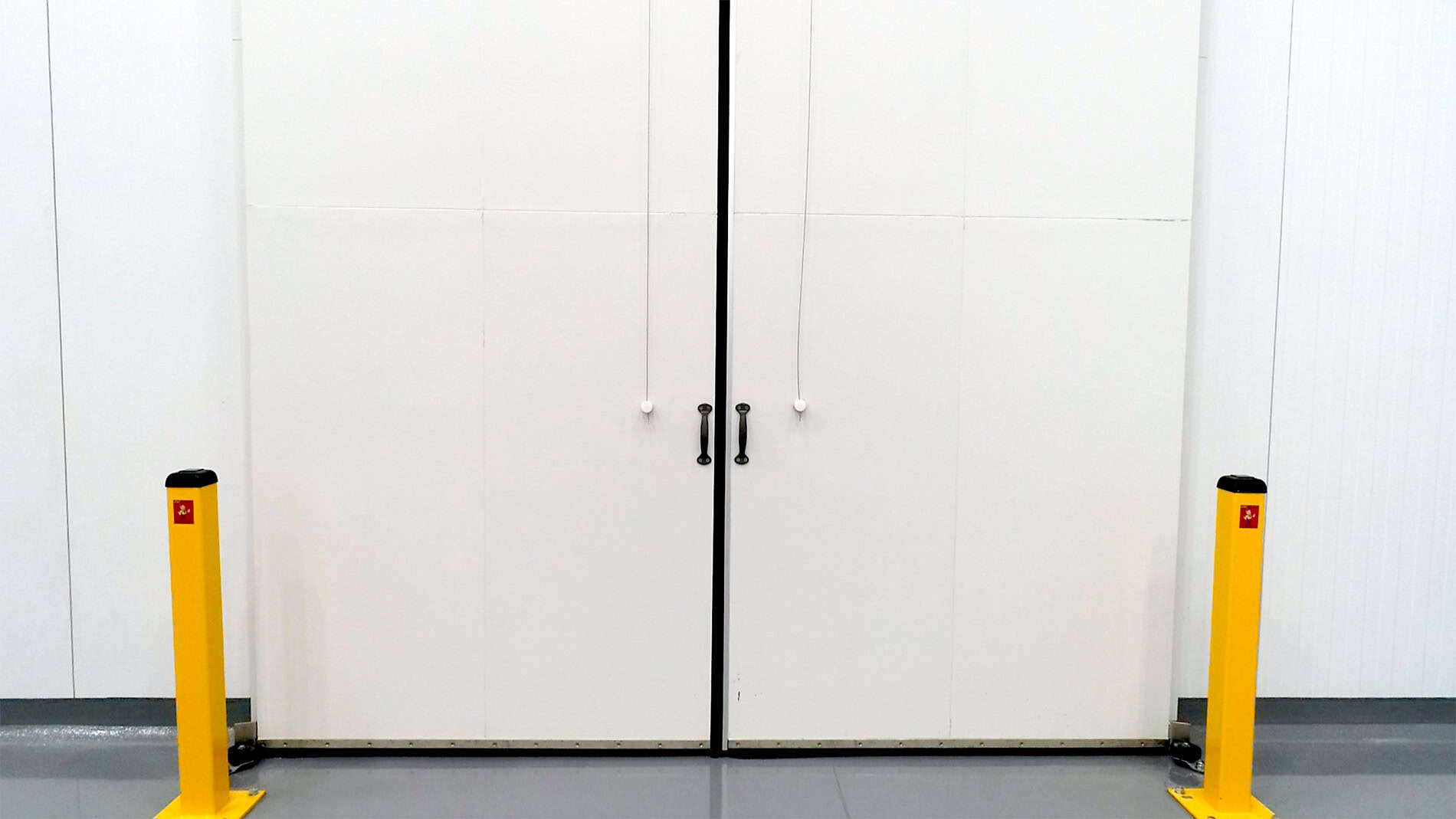 The front of a large cooler has two yellow safety bollards outside closed white metal doors.