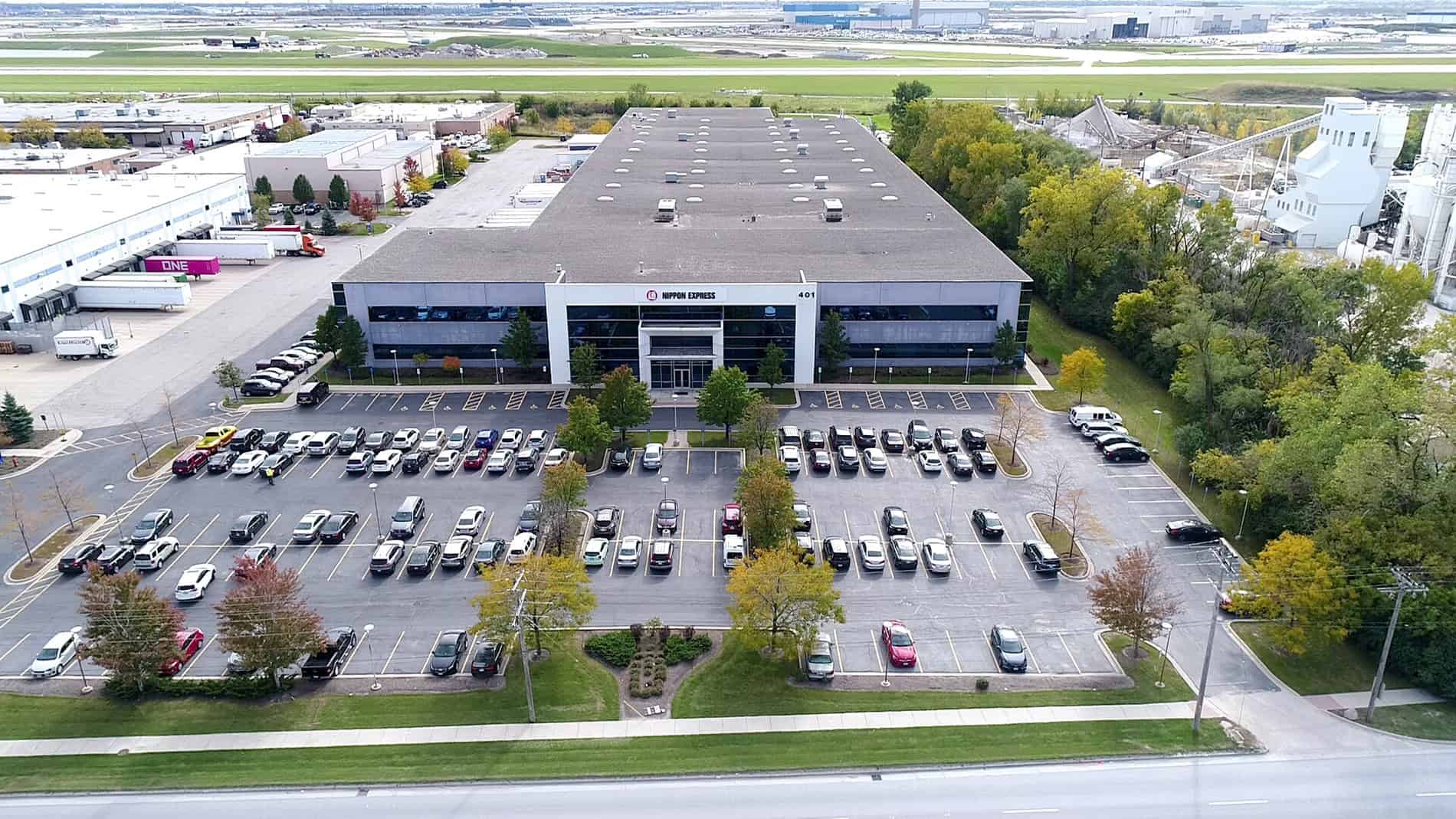 This is a distant bird' s-eye view of the entrance to Nippon Express and the front parking lot. The right side has a row of trees.