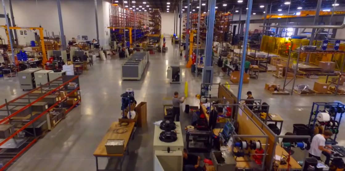 A second-floor view of a giant warehouse space, bustling with work, construction, and assembly amid tall metal shelves and workspaces.