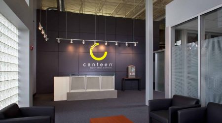 A welcome area with glass walls and comfortable seats. A reception area is in the back, and behind the desk is the word “Canteen” under their logo.