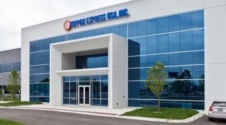 A white and grey office building behind a parking lot has blue mirror windows along the entrance wall and trees on each side of the front door. The sign above the entrance says “Nippon Express.”