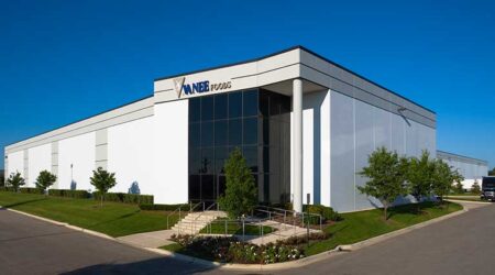 A large, white corporate office and warehouse facility stands against a clear blue sky. Steps and a wheelchair ramp lead to the front door. A logo consisting of a grey triangle and the words “Vanee Foods” are above the windowed front entrance.