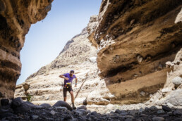 La imagen editorial captura a un excursionista caminando por un cañón desértico accidentado, rodeado de formaciones rocosas empinadas y estratificadas. Vestidos con una camiseta violeta y una mochila, caminan por las rocas iluminadas por el sol bajo un cielo azul claro, encarnando la aventura y la soledad.