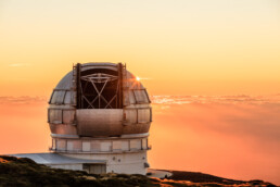 Un gran observatorio telescópico con estructura en forma de cúpula se alza majestuoso contra el impresionante cielo del atardecer, pintado en tonos naranjas y rosados. El horizonte y las nubes distantes se mezclan a la perfección, creando una escena serena y pintoresca.