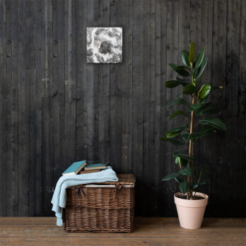Una canasta de mimbre llena de libros y una manta azul claro descansa sobre un piso de madera oscura contra una pared texturizada, capturando la esencia del estilo Arte Decorativo. Cerca, una planta en maceta con hojas verdes brillantes agrega vida a la escena, mientras que "Lienzo roca y sal", una pintura abstracta en blanco y negro de Volcanica Productions, cuelga encima.
